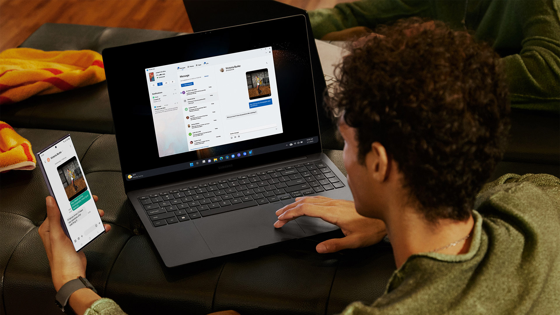 Uomo su divano scuro usa laptop e smartphone, entrambi con app di messaggistica. Sullo sfondo una coperta gialla.