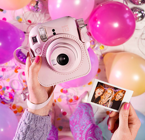 Mani tengono macchina fotografica rosa e foto di tre donne su sfondo con palloncini rosa e viola e coriandoli.