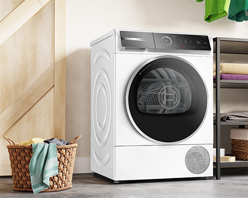 Asciugatrice bianca vicino a cesto di vimini e scaffale, su pavimento grigio. Panni appesi sullo sfondo.