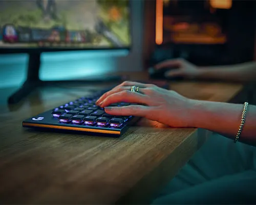 Mano su tastiera da gioco illuminata su scrivania di legno. Schermo acceso sullo sfondo.