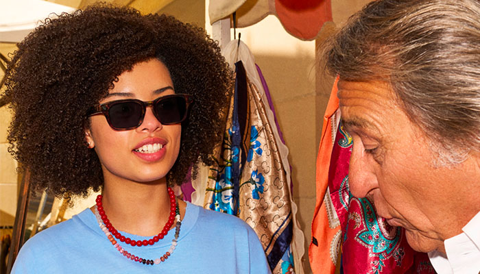 Una ragazza con capelli ricci afro, maglietta azzurra e occhiali da sole tartarugati sorride mentre parla con un uomo di profilo, in un mercato o negozio di abbigliamento.