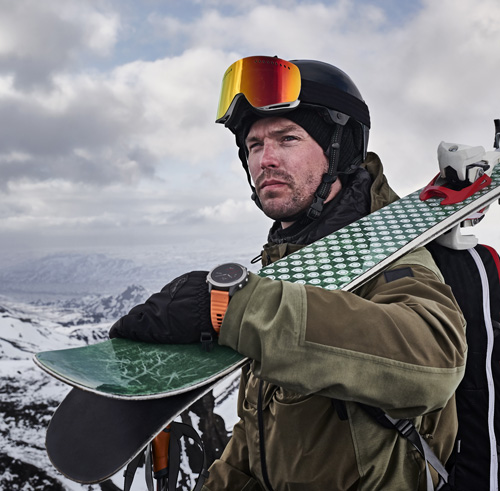 Scialpinista con casco, occhiali e sci sulla spalla. Indossa un orologio arancione. Nuvole e montagne innevate sullo sfondo.