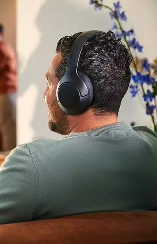 Uomo con cuffie nere seduto su un divano marrone. Sullo sfondo, fiori blu e una persona sfocata.