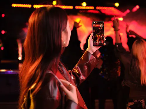 Donna che registra un evento con il telefono in una sala illuminata di rosso con una folla sullo sfondo.