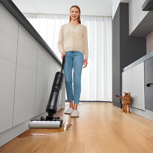 Donna pulisce il pavimento in legno con un aspirapolvere nero; un gatto arancione sta in piedi sullo sfondo.