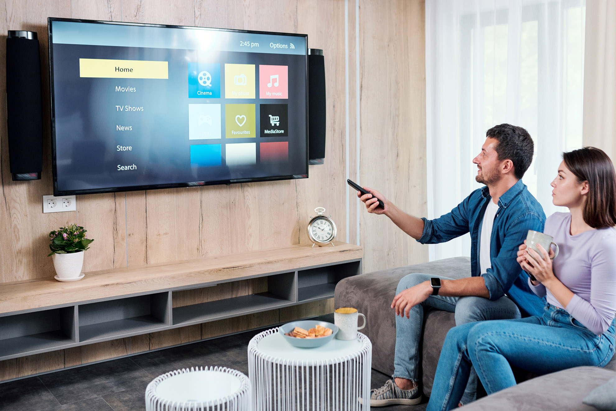 Soggiorno con TV a schermo piatto che mostra un menu, coppia seduta su un divano con bevande e telecomando.