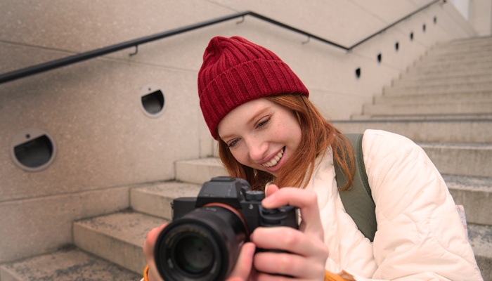 Una donna con i capelli rossi e un berretto bordeaux sorride mentre guarda lo schermo della fotocamera reflex che tiene tra le mani, seduta su una scalinata esterna.