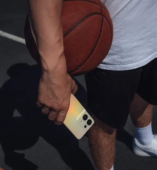 Primo piano di una persona che tiene un pallone da basket e un telefono color oro su un campo.