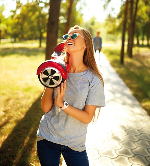 Una donna sorridente tiene in mano un hoverboard rosso e nero in un parco soleggiato. Un sentiero lastricato si estende sullo sfondo.