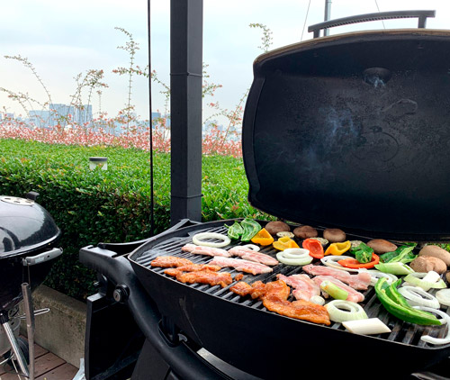 Griglia con carne e verdure su terrazza. Sullo sfondo, vegetazione e edifici urbani.