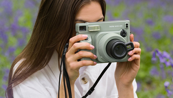 Donna con camicia bianca che tiene in mano una fotocamera verde. Fiori viola sullo sfondo.