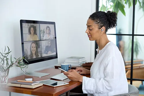 Donna a una scrivania con un computer che mostra una videochiamata. Ci sono libri, un tablet, una tastiera e una tazza.