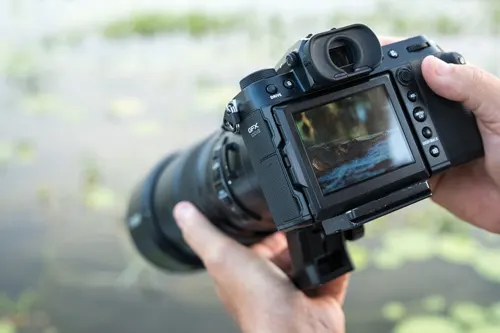 Cameraman che tiene in mano una fotocamera nera con obiettivo, con acqua sullo sfondo.