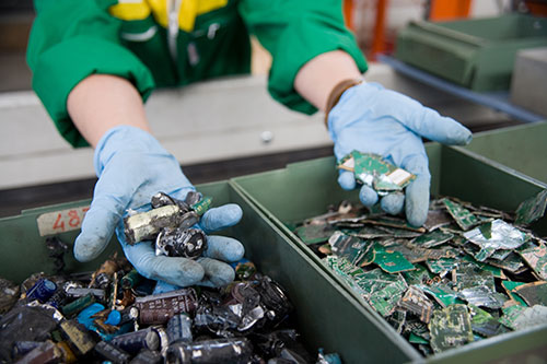 Mani guantate selezionano scarti elettronici da contenitori verdi. Uno contiene schede verdi, l'altro componenti misti scuri.
