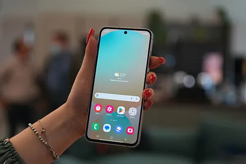 Mano che tiene un telefono con sfondo azzurro e icone colorate. Sullo sfondo, persone e luci soffuse.