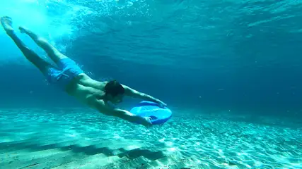 Uomo in costume da bagno blu che nuota sott'acqua. Tiene una tavola blu. Il fondale marino è visibile.