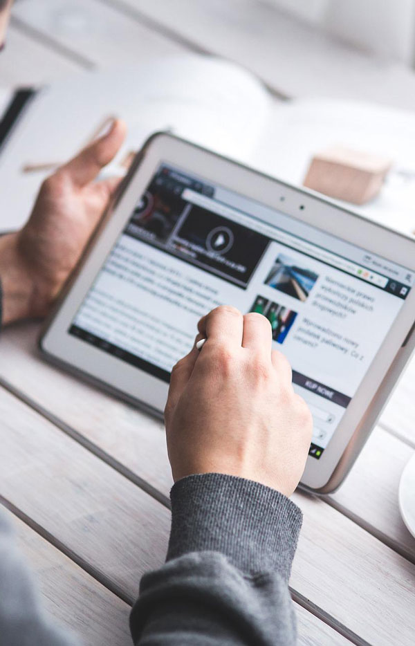 Mani che tengono un tablet su un tavolo di legno bianco. Sullo schermo del tablet si vede un sito web con video e testo.