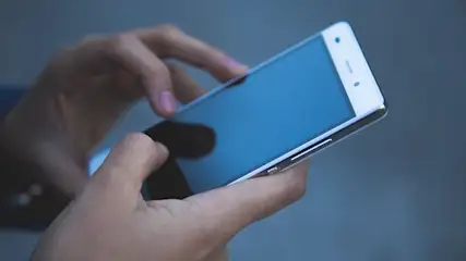 Mani che tengono un telefono bianco con schermo blu. Un dito tocca lo schermo, su sfondo blu uniforme.