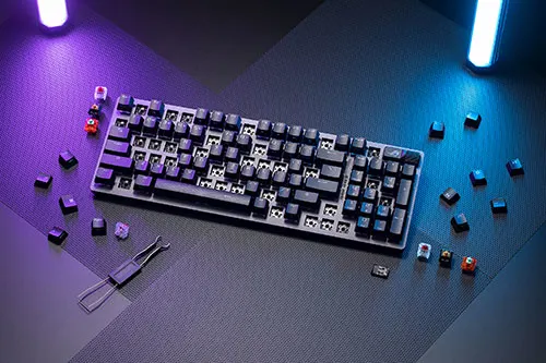 Tastiera nera con alcuni tasti rimossi, pinzette e luci blu e viola su una superficie scura.
