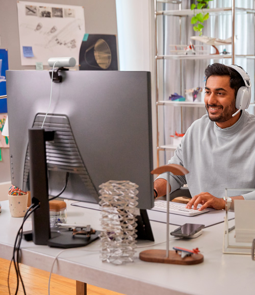 Uomo con cuffie al computer su scrivania bianca con oggetti, scaffali sullo sfondo.