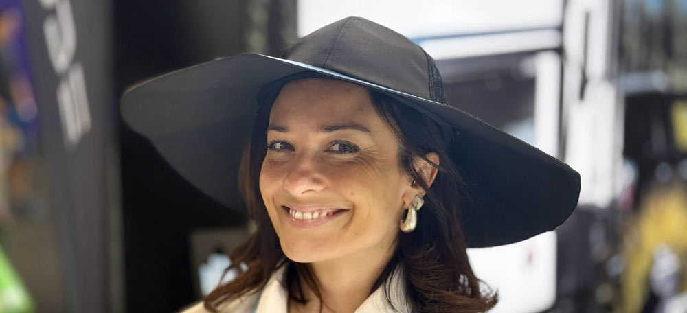 Donna sorridente con cappello nero a tesa larga e orecchini pendenti, indossa camicia bianca. Sfondo sfocato.
