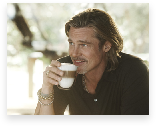 Uomo con camicia scura che beve da una tazza bianca a strati. Indossa bracciali e una collana. Sfondo sfocato di alberi e luce solare.