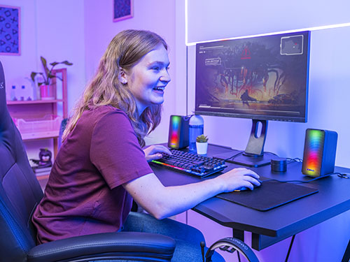 Ragazza seduta su una sedia da gaming, usa tastiera e mouse di fronte a un monitor. Ci sono altoparlanti illuminati e scaffali sullo sfondo.