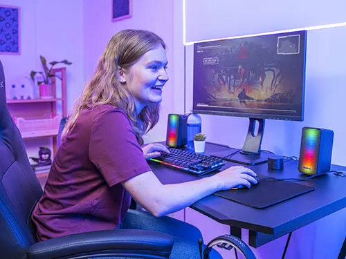 Ragazza seduta su una sedia da gaming, usa tastiera e mouse di fronte a un monitor. Ci sono altoparlanti illuminati e scaffali sullo sfondo.