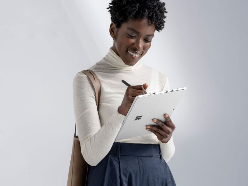 Donna sorridente usa una penna su un tablet bianco. Indossa un dolcevita bianco, una gonna blu, con una borsa a tracolla marrone. Lo sfondo � grigio.