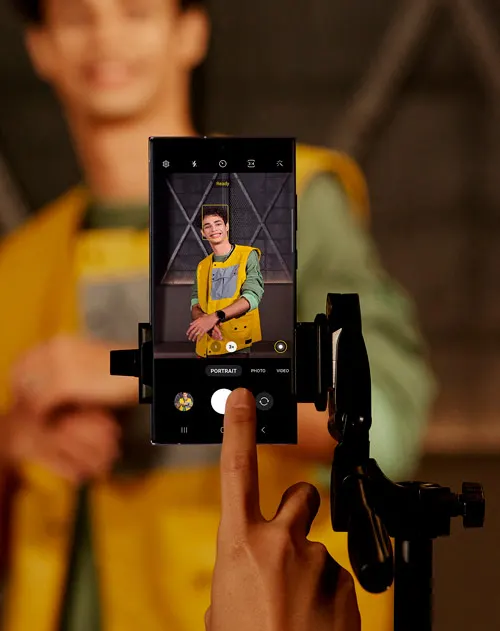 Primo piano di dito su schermo smartphone su treppiede. Schermo mostra uomo con gilet giallo. Persona sfocata sullo sfondo.