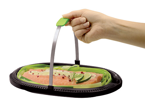 Mano che solleva una vaporiera nera ovale con salmone, lime e erbe aromatiche su un letto di verdure verdi.