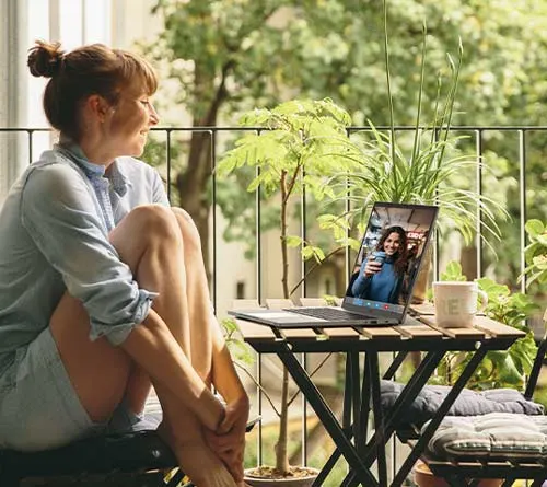 Donna seduta su un balcone con un laptop su un tavolino tra piante e ringhiera in ferro. Sul laptop una videochiamata.