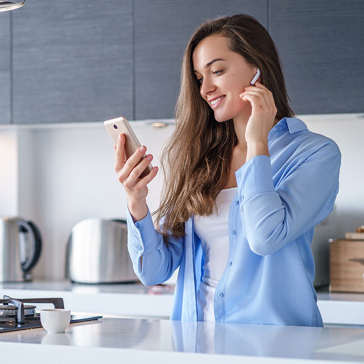 Donna sorride, indossa auricolari e camicia azzurra, guarda un telefono. Sul bancone ci sono tazza, bollitore e tostapane.