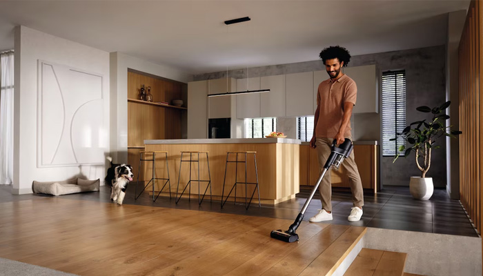 Un uomo aspira un pavimento in legno. Un cane è in piedi sullo sfondo. C'è una cucina con isola e sgabelli.