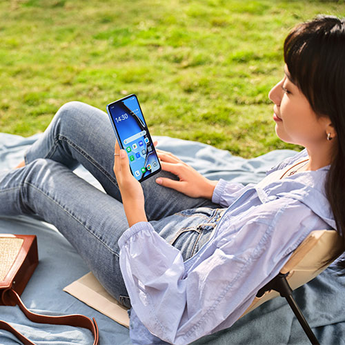 Donna su prato, seduta su sedia, con smartphone in mano che mostra l'ora, su coperta blu, accanto a borsa e libro.