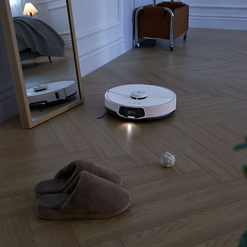 Aspirapolvere robot bianco sul pavimento in legno, vicino a pantofole marroni e carta appallottolata. Specchio che riflette la scena.