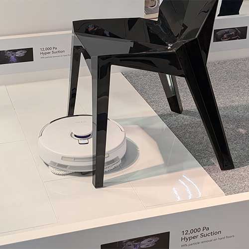 Aspirapolvere robot bianco sotto una sedia nera su una superficie bianca. Sullo sfondo si vede moquette grigia.