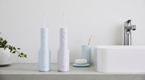 Due irrigatori orali bianchi su un ripiano, vicino a un lavandino, un rubinetto, un sapone e una pianta in vaso.