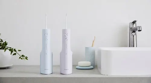 Due irrigatori orali bianchi su un ripiano, vicino a un lavandino, un rubinetto, un sapone e una pianta in vaso.