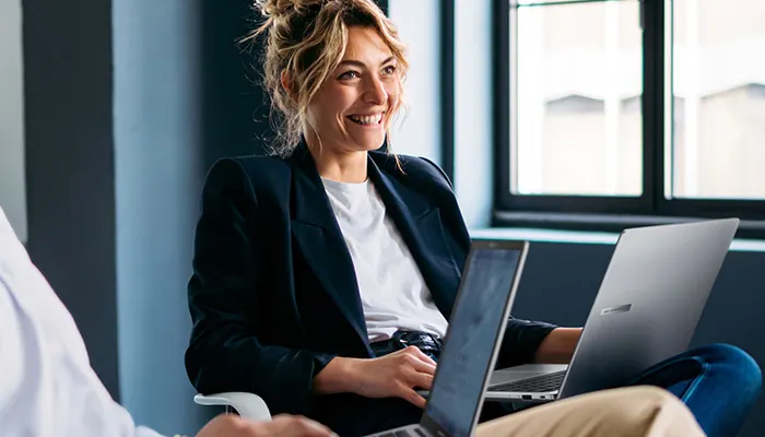 Donna sorridente con blazer blu e maglietta bianca, seduta con due laptop. Finestra sullo sfondo.