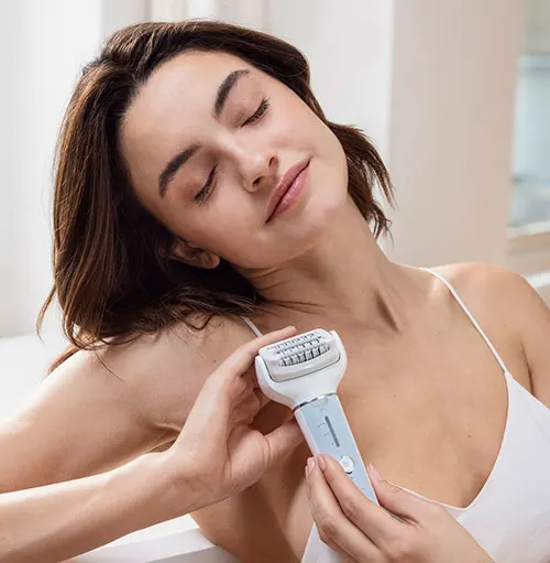 Donna con occhi chiusi tiene in mano un epilatore bianco e azzurro. Indossa una canottiera bianca.