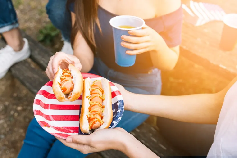 ragazze mangiano degli hotdog sedute su una panchina con piatti decorati con la bandiera americana.