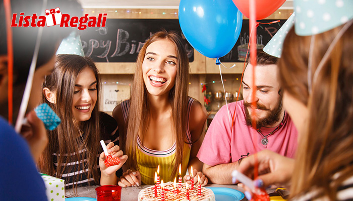 Festa di compleanno con torta, candele accese e palloncini colorati. Persone sorridenti indossano cappellini.