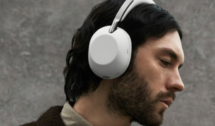 Inquadratura di profilo di un uomo con capelli scuri e barba che indossa grandi cuffie over-ear bianche, con uno sfondo grigio materico.