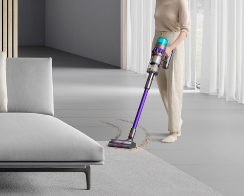 Donna aspira la polvere sul tappeto grigio con un aspirapolvere viola e argento. Divano grigio sullo sfondo.