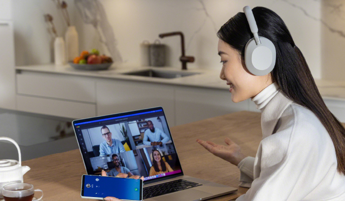 Donna con cuffie che partecipa a una videochiamata su un laptop. Un telefono � appoggiato accanto al laptop. Sullo sfondo si vede una cucina.