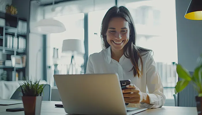 Donna sorridente usa il telefono davanti al laptop. Sul tavolo ci sono piante e un altro telefono. Dietro, libreria e lampada.