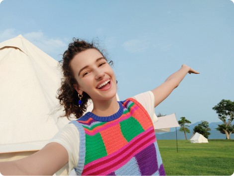 Donna sorridente in gilet a righe, braccio alzato. Dietro, tenda bianca su prato verde sotto cielo azzurro.