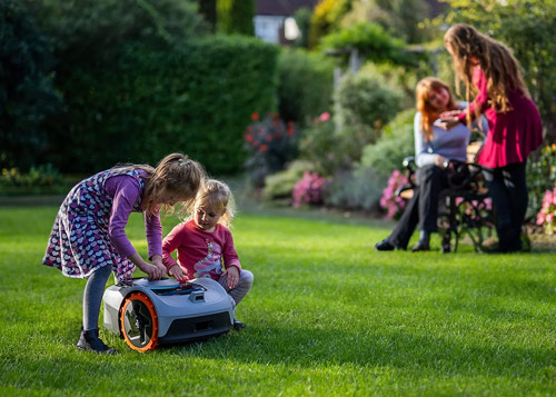 Due bambine interagiscono con un robot tagliaerba su un prato verde. Dietro, due donne sedute su una panchina in un giardino.