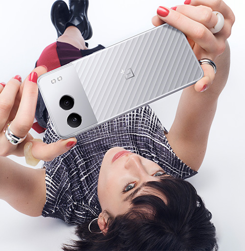 Donna sdraiata su sfondo bianco, tiene un telefono argento con doppia fotocamera. Indossa una camicia a quadri e scarpe nere.